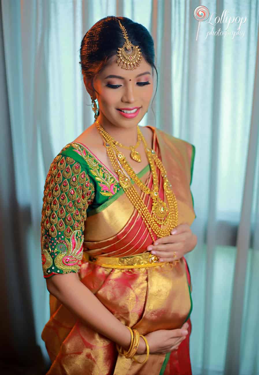 An expectant mother stands glowing in traditional South Indian attire, adorned with exquisite gold jewelry, captured in a soft, illuminated indoor setting in Chennai.