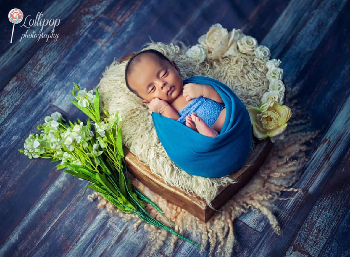 Newborn sleeping in a cozy blue wrap, surrounded by a rustic floral setup, blending softness and nature in Chennai.